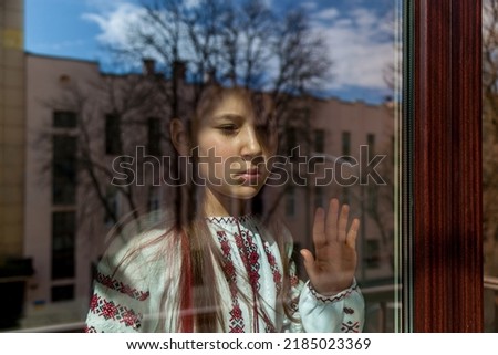 A girl with a sad face in an embroidered dress stands near the window. The girl protests against the war, and shows patriotism and love for Ukraine