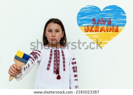 A girl in an embroidered dress stands against a white wall, holding a brush painted blue and yellow. The girl protests against the war.Ukrainian-Russian conflict, war in Ukraine