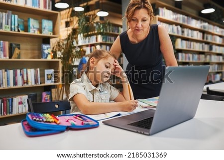 Teacher helping to solve and finish task. Bored little student girl doing her homework using laptop in after school club at primary school. Back to school. Child using technology on elementary class