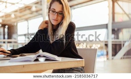 Caucasian young female student with blond hair wearing eyeglasses and reading books on table in college library