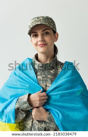 Portrait of smiling soldier covering with ukraining flag isolated on grey