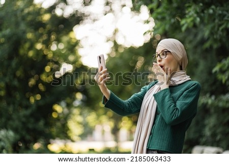 Pretty smiling muslim business woman in hijab walks around the park and using smartphone during video chat. Happy arabic girl standing outdoor and checking email on digital gadget. Lifestyle concept.