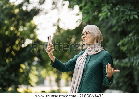 Pretty smiling muslim business woman in hijab walks around the park and using smartphone during video chat. Happy arabic girl standing outdoor and checking email on digital gadget. Lifestyle concept.
