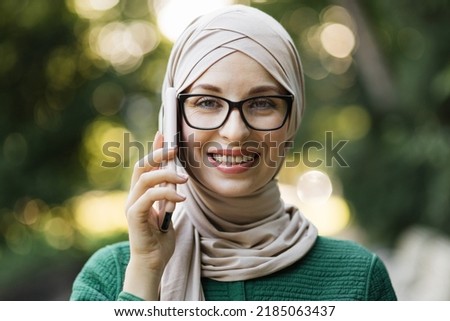 Modern stylish muslim woman in hijab and casual clothes walking in city park. Arab female talking on smartphone, taking selfie, or doing video call outdoor.