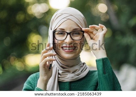 Modern stylish muslim woman in hijab and casual clothes walking in city park. Arab female talking on smartphone, taking selfie, or doing video call outdoor.
