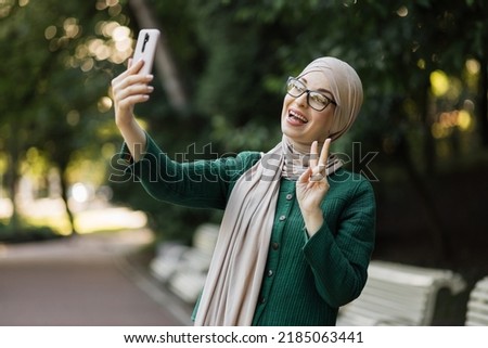 Portrait of smiling happy beautiful muslim woman relaxing using digital smartphone showing thumb up. Young muslim girl looking at screen speaking during online video call on social media on city.