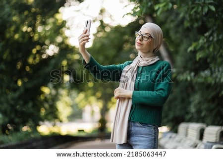 Young Muslim Arab woman in hijab takes a selfie of herself with the city background. Elegant, tall, slim and wearing casual clothes and turban, smiling while takes her photo.