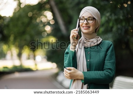 Portrait of smiling young muslim woman doing a phone call, taking on additional work during the break. Cheerful arabic woman in hijab using smart phone outdoor walking in park.