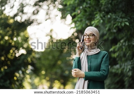 Portrait of smiling young muslim woman doing a phone call, taking on additional work during the break. Cheerful arabic woman in hijab using smart phone outdoor walking in park.