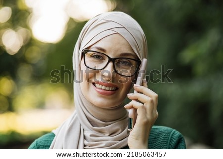 Cheerful young islamic businesswoman talking on mobile phone while walking on city park. Beautiful young woman with hijab working using smartphone during break.