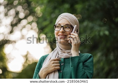 Cheerful young islamic businesswoman talking on mobile phone while walking on city park. Beautiful young woman with hijab working using smartphone during break.