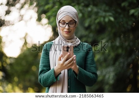 Attractive smiling muslim woman in hijab texting on smart phone standing on background of green city park. Portrait of young arabian businesswoman using mobile phone outdoor.