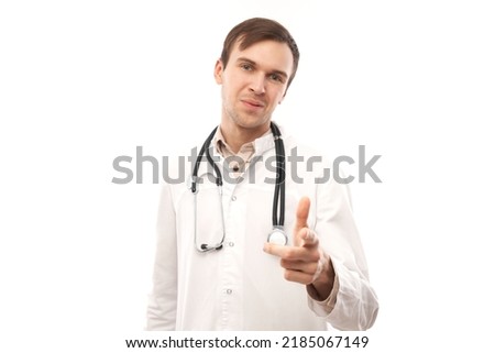 Portrait of friendly positive young male doctor in white medical coat chooses you, points finger at camera isolated on white background
