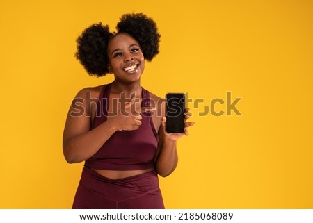 Smiling, happy, young girl with afro hairstyle posing on yellow studio background in sporty clothes, holding mobile phone in one hand showing new fitness app. Sale concept. Copy space.
