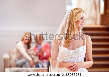 Grandmother With Mother Watching Adult Daughter Choosing And Trying On Wedding Dress In Bridal Store