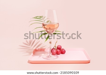Rose wine. Minimalistic, Summer, holiday composition with glass of rose wine, grapes and palm leaf on a pink tray on a pink background. Self-partnered and self love concept