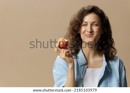 Funny happy cute curly beautiful female in jeans casual shirt chewing enjoy apple posing isolated on over beige pastel background. Healthy food. Natural eco-friendly products concept. Copy space Royalty-Free Stock Photo #2185103979