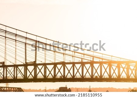 People cross a suspension bridge at sunset. Silhouette profile shot of bridge with low tree-line and bridge in background. 