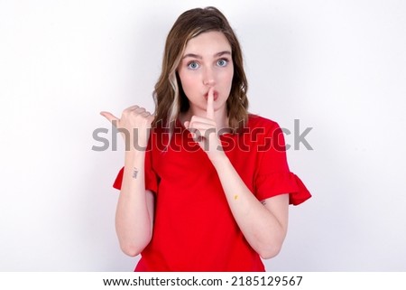 young caucasian woman wearing red T-shirt over white background asking to be quiet with finger on lips pointing with hand to the side. Silence and secret concept.