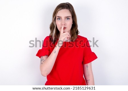 young caucasian woman wearing red T-shirt over white background  makes silence gesture, keeps finger over lips. Silence and secret concept.