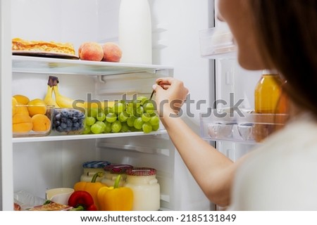 Woman hand taking, grabbing or picks up green bunch of grapes out of open refrigerator shelf or fridge drawer full of fruits, vegetables, banana, peaches, yogurt. Healthy food diet, lifestyle Royalty-Free Stock Photo #2185131849