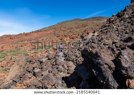 Woman hiking on field of solidified lava, ash and pumice on volcanic terrain. Dark rocks on bare terrain. Trail to summit volcano Pico del Teide, National Park, Tenerife, Canary Islands, Spain, Europe Royalty-Free Stock Photo #2185140041