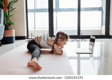 Pretty caucasian little girl in sportswear watching online video on laptop and doing fitness exercises at home. Distant training with personal trainer, social distance or self-isolation, online