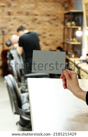female hand holding grey postcard or bossiness card with mock up on stylish barbershop or beauty salon background. blurred barber man is cutting and shaving the beard o client