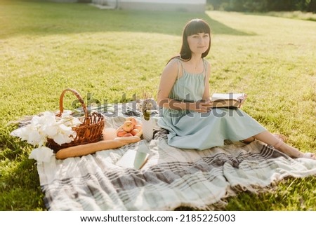 A beautiful young European woman is sitting on a blanket and holding a book to read. A girl on a picnic in a summer park. High quality photo