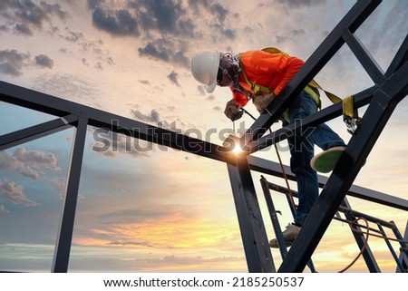 A welder is welding steel on a steel roof truss. Working at height equipment. Fall arrestor device for worker with hooks for safety body harness. Worker in construction site. Royalty-Free Stock Photo #2185250537