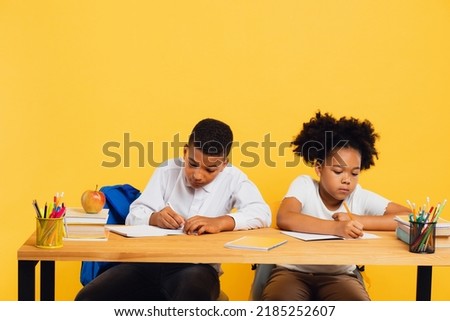 Happy african american schoolgirl and mixed race schoolboy sitting together at desk and studying on yellow background. Back to school concept. Royalty-Free Stock Photo #2185252607