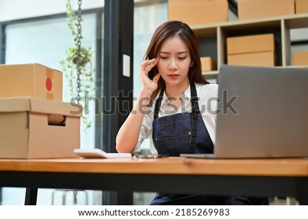 Stressed young Asian female online startup business working at her office desk in stock room, worried about her sales.