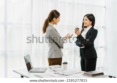Two young friends Asian business women standing and discussing near the window in the office workplace.