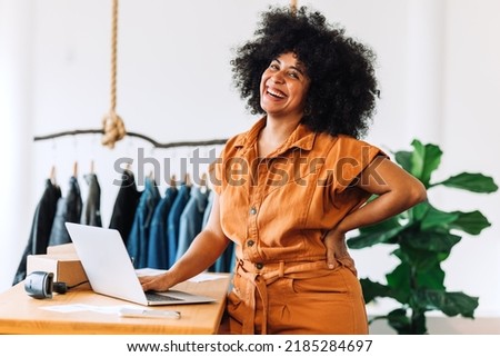Black small business owner smiling at the camera while standing in her shop. Happy businesswoman managing her clothing orders on a laptop. Young female entrepreneur running an online clothing store.