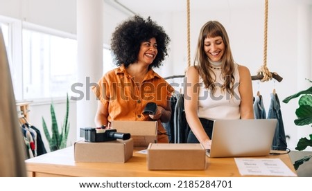 Cheerful clothing store owners preparing online orders for shipping. Two happy businesswomen smiling while working in a thrift store. Female entrepreneurs running an e-commerce small business. Royalty-Free Stock Photo #2185284701