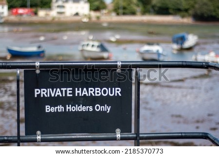 Private harbour berth holders only sign in black at a pretty harbor at low tide. Softly focused boats sitting on the mud on the background contrast the sharply focused black warning sign and railings