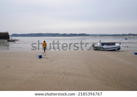Woman in bright yellow jacket walking on a sandy beach towards the sea at low tide, with small boats moored in the harbor Royalty-Free Stock Photo #2185396767