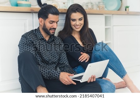Woman with long dark hair smiling looking at laptop on her boyfriend's lap sitting on the floor together by kitchen counter. Young couple. Indoor shot. High quality photo