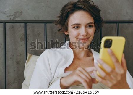 Close up young woman in white shirt pajama she lying in bed hold use mobile cell phone rest relax spend time in bedroom lounge home in own room hotel wake up dream be lost in reverie good mood day. Royalty-Free Stock Photo #2185415569
