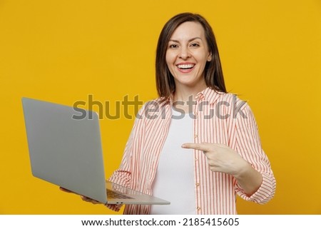 Young happy smiling woman she 30s in striped shirt white t-shirt hold use work point index finger on laptop pc computer isolated on plain yellow background studio portrait. People lifestyle concept