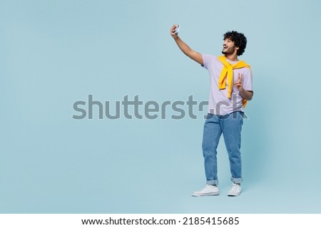 Full size young bearded Indian man 20s years old wears white t-shirt doing selfie shot on mobile cell phone post photo on social network isolated on plain pastel light blue background studio portrait