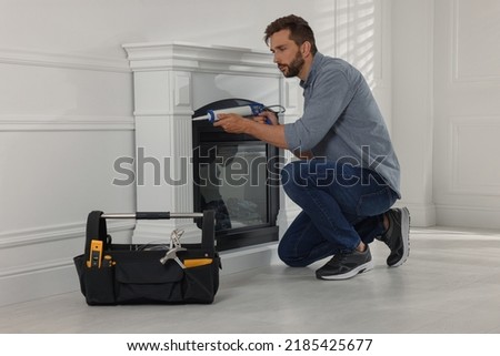 Man sealing electric fireplace with caulk near white wall in room Royalty-Free Stock Photo #2185425677