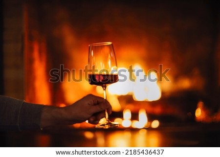 Man having red wine by the fireplace. Close up of man holding and drinking red wine glass by open wood fire place relaxing and enjoying quiet moment at home in winter holiday vacation, Christmas 