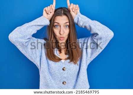 Young woman standing over blue background doing funny gesture with finger over head as bull horns 