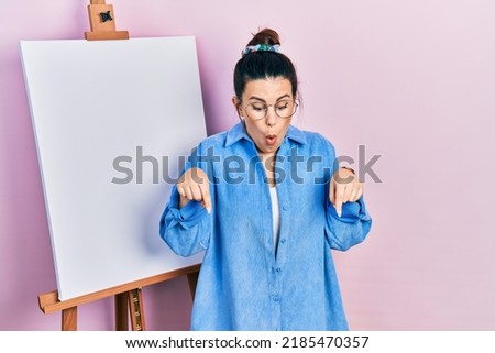 Young hispanic woman standing by painter easel stand pointing down with fingers showing advertisement, surprised face and open mouth 
