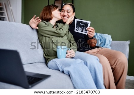 Portrait of kissing gay couple expecting baby and showing ultrasound image to family via video chat, copy space
