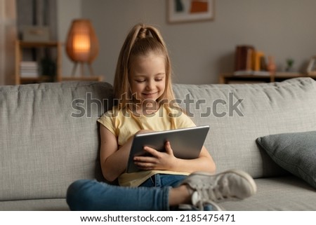Happy Kid Girl Using Digital Tablet Browsing Internet Sitting On Couch At Home. Child Having Fun Playing Online Game On Computer In Living Room Indoor. Children And Gadgets Concept. Selective Focus