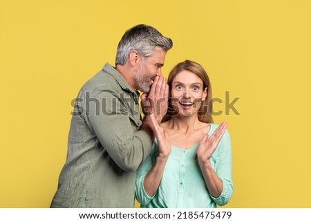 Middle aged man sharing secret or whispering gossips into his wife's ear on yellow studio background. Caucasian spouses sharing shocking news, discussing rumors together