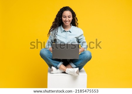 Smiling Arabic Female Freelancer Using Laptop Computer Working Remotely Online Sitting On Cube In Studio Over Yellow Background. Freelance Career And Technology Concept