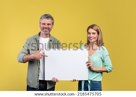 Place for your ad. Happy mature spouses holding empty blank placard board and smiling at camera, man pointing at white paper over yellow studio background Royalty-Free Stock Photo #2185475705